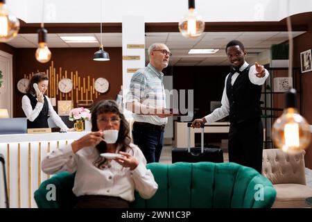 Sympathique chasseur afro-américain assiste un homme senior à la réception de l'hôtel avec des bagages dans la chambre réservée. Une femme âgée visiteuse sirotant du thé en attendant l'enregistrement dans le salon confortable avec canapés. Banque D'Images