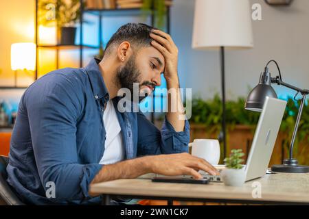 Ennuyé jeune homme barbu indien endormi travaillant sur un ordinateur portable, penché sur la main au bureau à domicile. Épuisé homme arabe fatigué freelance homme bourré de travail. Emploi, profession, sans travail. Banque D'Images