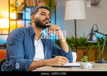 Homme barbu indien freelance travaillant avec des documents du bureau à domicile, analysant des informations, lisant des documents d'affaires préparent le rapport financier. Jeune comptable arabe analysant des documents graphiques. Banque D'Images
