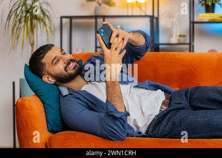 Sourire bel homme indien prenant des écouteurs sans fil hors de la boîte, placer des gadgets dans les oreilles, démarrer le lecteur multimédia à l'aide d'un smartphone, écouter de la musique, profiter des loisirs, se détendre sur le canapé à la maison. Banque D'Images