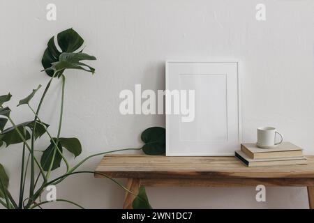 Maquettes de cadre photo vertical sur banc en bois vintage, table. Tasse de café, thé sur pile de livres. Usine de fromage en pot vert Monstera. Mur blanc Banque D'Images