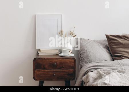 Maquette de cadre photo blanc vertical sur de vieux livres. Vue élégante sur la chambre. Oreillers marron, gris lin et velours, couverture. Table de nuit en bois avec céramique Banque D'Images