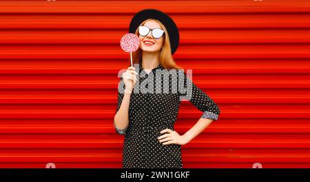 Portrait d'été de jeune femme souriante heureuse avec sucette colorée portant un chapeau rond noir sur fond rouge Banque D'Images