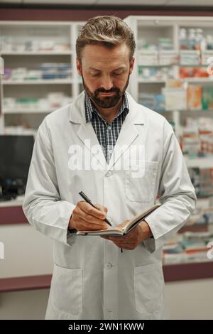 Le pharmacien masculin concentré en uniforme prend des notes dans le bloc-notes lorsqu'il travaille en pharmacie Banque D'Images