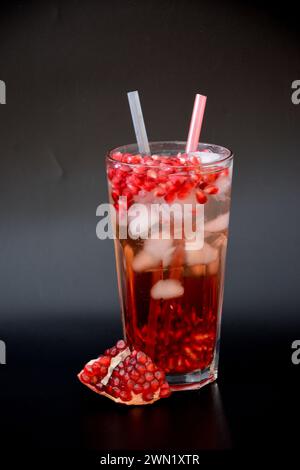 Un grand verre à facettes de jus de grenade avec de la glace et des graines sur un fond noir, à côté d'un morceau de fruit mûr. Disposition verticale. Banque D'Images