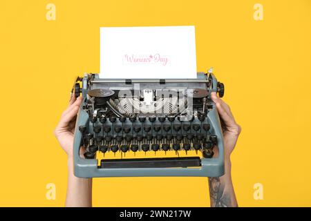 Mains tenant la machine à écrire vintage et la carte postale festive avec le texte HAPPY WOMEN'S DAY sur fond jaune Banque D'Images
