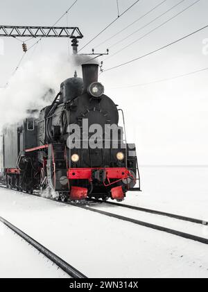 Vieille locomotive à vapeur circulant dans un champ enneigé. Locomotive vintage noire tirant un train un jour d'hiver. Banque D'Images