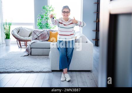 Photo du corps entier de charmante belle vieille dame assis canapé tenir la clé de maison démontrer l'approbation pouce vers le haut intérieur spacieux à l'intérieur Banque D'Images