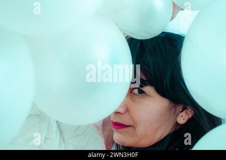 Gros plan du visage d'une femme latino entourée de ballons blancs regardant la caméra, performance artistique, danse ludique Banque D'Images