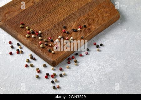 Planche à découper beige en bois avec des pois de poivre multicolores dispersés sur un comptoir en pierre grise. Banque D'Images