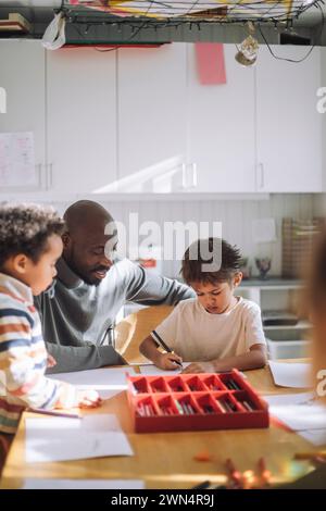 Enseignant masculin aidant garçon tout en écrivant sur papier au banc dans la salle de classe Banque D'Images