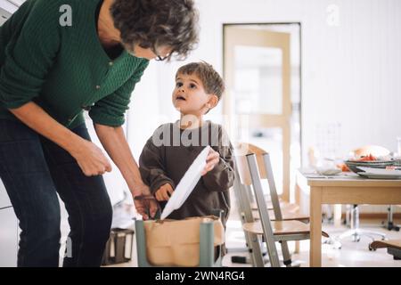 Garçon regardant une enseignante tout en jetant de la nourriture dans la poubelle à la maternelle Banque D'Images