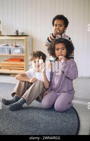 Portrait de garçons et de filles mangeant de la crème glacée assis sur le tapis dans la salle de classe à la maternelle Banque D'Images