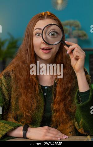 Enquêteur excité femme caucasienne étonnée tenant une loupe près du visage, regardant dans la caméra avec un gros œil drôle zoomé, wow, la recherche, l'analyse. Fille au lieu de travail de table de bureau à domicile. Vertical Banque D'Images