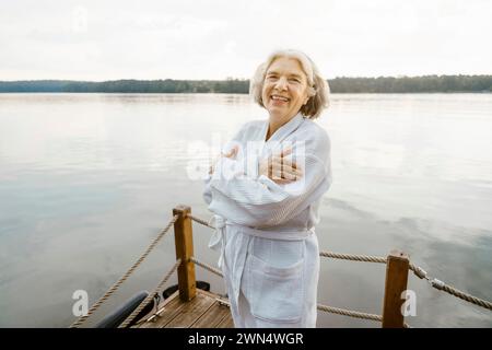 Portrait d'une femme âgée heureuse portant un peignoir blanc s'embrassant sur la péniche près de la rivière contre le ciel Banque D'Images