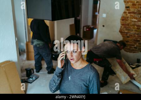 Femme responsable de la construction parlant sur le téléphone portable sur le site tandis que les travailleurs travaillent en arrière-plan Banque D'Images