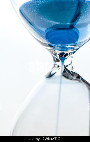 Une vue rapprochée de sable bleu coulant à travers la taille étroite d'un sablier sur un fond blanc avec l'accent sur le point central où le sable Banque D'Images