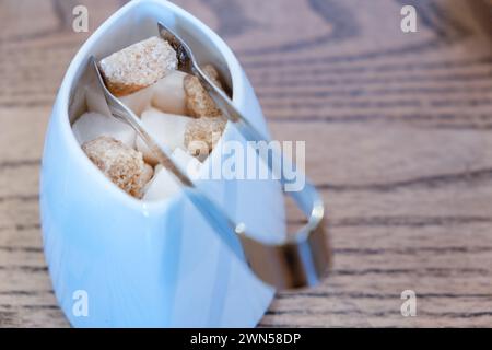 Une petite paire de pinces à sucre saisissant un cube de cassonade dans un petit bol à sucre. Le sucre est fourni dans un café pour sucrer le thé et les boissons au café Banque D'Images