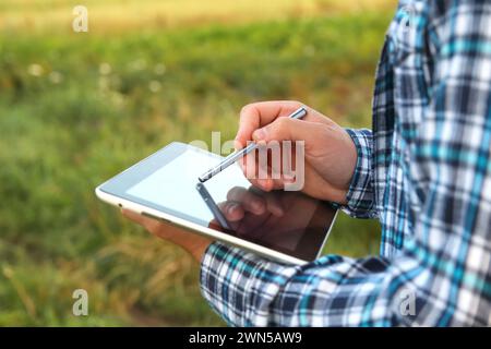 Un agriculteur agronome utilise une tablette numérique dans un jeune champ de maïs au lever ou au coucher du soleil. Gros plan des mains avec l'écran de la tablette et le stylet. Banque D'Images