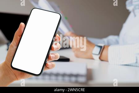 Maquette écran blanc de smartphone dans la main féminine devant la femme d'affaires travaillant au bureau. Banque D'Images