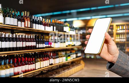 Main féminine montrant l'écran vide de son téléphone intelligent devant le magasin d'alcool avec des bouteilles de vin sur les étagères. Banque D'Images