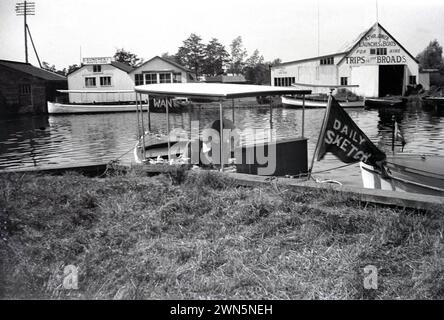 Années 1950, historique, chantiers navals sur le Norfolk large....vu sur la droite est le chantier naval d'Un Thrower, offrant des lancements et des bateaux à louer etTrips to the Broads. Au premier plan, un homme sur un petit bateau, avec un drapeau disant, Daily Sketch, un journal tabloïd national de l'époque. Banque D'Images