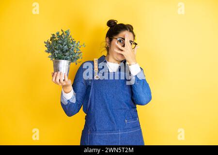 Jeune femme jardinière caucasienne tenant une plante isolée sur fond jaune jetant un coup d'œil dans le choc couvrant le visage et les yeux avec la main, regardant à travers le doigt Banque D'Images