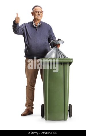 Homme mûr souriant jetant un sac à déchets dans une poubelle et faisant des gestes avec les pouces isolés sur fond blanc Banque D'Images