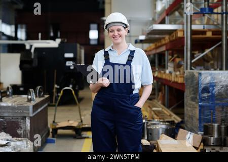 Plan moyen long d'une jeune femme caucasienne joyeuse ouvrière tenant un presse-papiers souriant à la caméra Banque D'Images