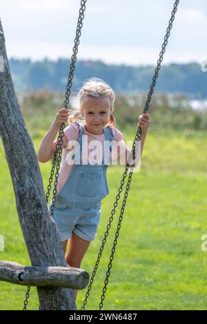 Une fille en salopette en denim saisit les chaînes d'une balançoire, prête pour le mouvement ludique dans un parc ensoleillé, parfait pour les thèmes des enfants actifs et du jeu en plein air, et lui Banque D'Images