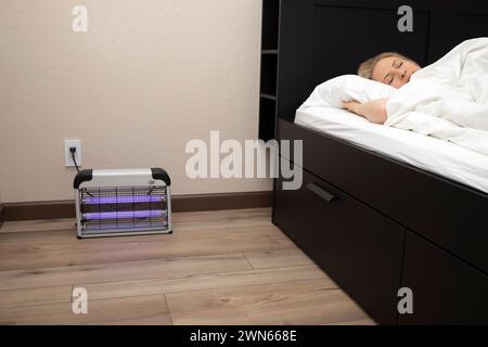 Moustique électrique ou anti-insectes avec lumières bleues violettes allumées près de la personne dormant dans le lit dans la chambre. Lampe anti-insectes sur plancher en bois dans la chambre Banque D'Images