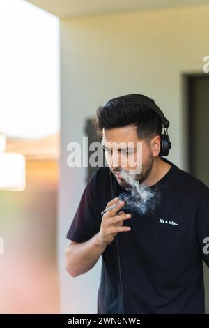 Jeune homme en vêtements noirs écoutant de la musique avec des écouteurs tout en fumant du cannabis Banque D'Images