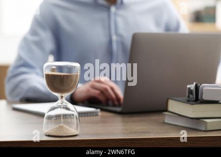 Sablier avec du sable coulant sur le bureau. Homme utilisant un ordinateur portable à l'intérieur, mise au point sélective Banque D'Images