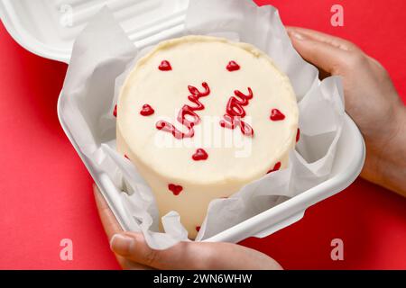 Femme tenant une boîte à emporter avec un gâteau au bento à la table rouge, gros plan. Prog Surprise pour la Saint-Valentin Banque D'Images