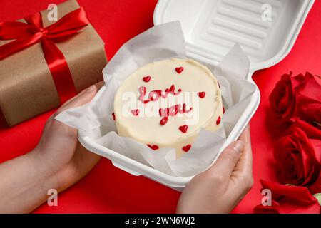 Femme tenant une boîte à emporter avec un gâteau au bento à la table rouge, gros plan. Prog Surprise pour la Saint-Valentin Banque D'Images