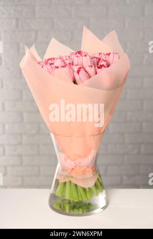 Bouquet de belles tulipes roses dans un vase sur une table blanche près du mur de briques Banque D'Images