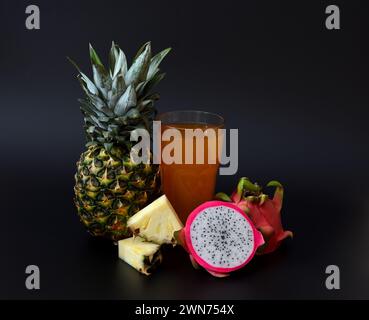 Un grand verre à facettes d’un mélange de fruits exotiques sur fond noir, à côté de morceaux d’ananas mûr et d’un fruit pitaya coupé. Gros plan. Banque D'Images