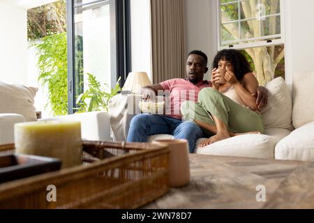 Couple afro-américain biracial assis près d'un canapé, regardant la télévision à la maison Banque D'Images