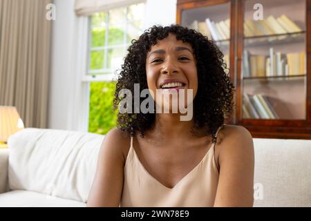 Jeune femme biraciale aux cheveux bouclés sourit chaleureusement lors d'un appel vidéo, assise sur un canapé blanc Banque D'Images