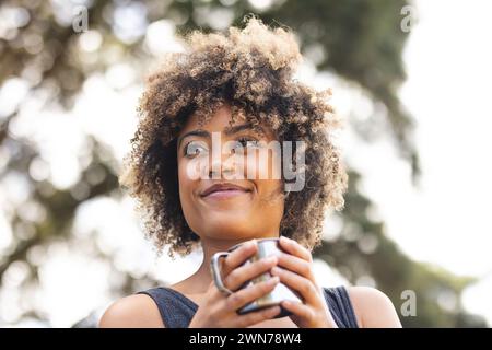 Une jeune femme biraciale aux cheveux bouclés tient une tasse à l'extérieur Banque D'Images