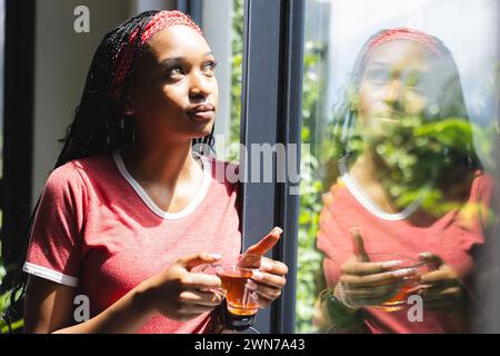 Une jeune femme afro-américaine regarde par une fenêtre, tenant une tasse de thé Banque D'Images