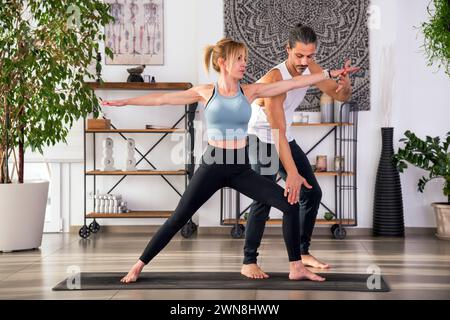 Concentrée entraîneur de yoga aidant la jeune femme à exécuter Virabhadrasana pendant la formation dans un studio de fitness moderne avec des étagères de plantes en pot an Banque D'Images