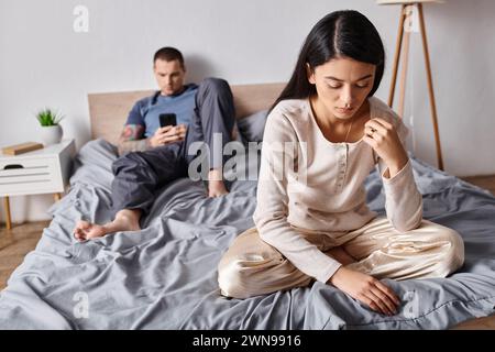 femme asiatique offensée assise près de son mari à l'aide d'un smartphone dans la chambre à coucher à la maison, conflit familial Banque D'Images