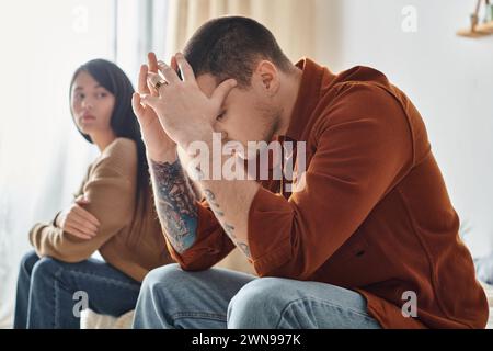 jeune homme bouleversé tatoué assis avec la tête inclinée près de la jeune femme asiatique, concept de divorce familial Banque D'Images