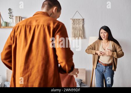 femme asiatique irritée se querellant contre le mari debout avec des vêtements dans la chambre, conflit familial Banque D'Images