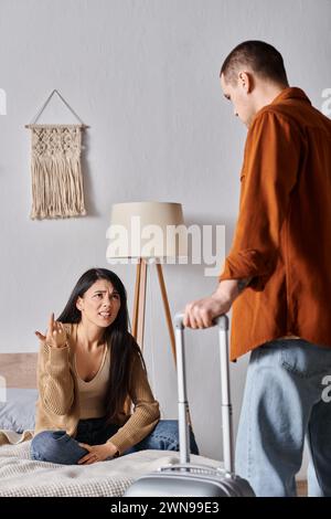 femme asiatique irritée assise sur le lit et se disputant avec le mari avec la valise à la maison, divorce Banque D'Images