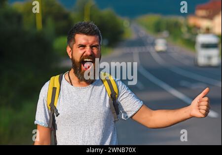 Arrêt automatique du déplacement. Taille basse essayez d'arrêter la voiture et le pouce vers le haut geste. Homme au visage joyeux et à la barbe voyageant en randonnée avec la route en arrière-plan Banque D'Images