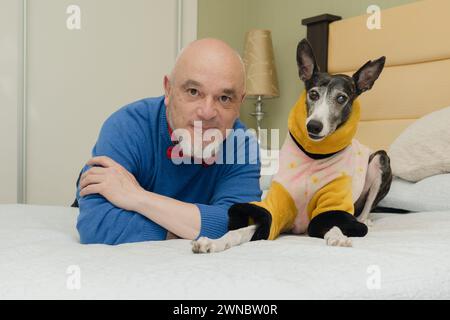 Portrait d'un homme d'âge moyen et de son chien lévrier vêtu de vêtements chauds, allongé sur le lit, regardant la caméra. Banque D'Images