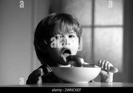 Jeune enfant assis sur la table mangeant avec une expression drôle sur le visage. Délicieux petit déjeuner pour les enfants. Délicieux petit déjeuner pour les enfants. Bébé gai enfant mange la nourriture lui-même Banque D'Images