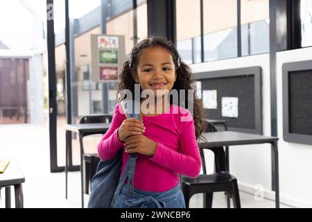 Fille biraciale aux cheveux bouclés souriant, tenant un sac à dos dans une classe d'école Banque D'Images